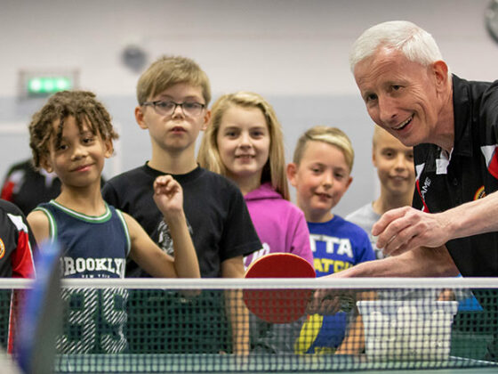 A table tennis coach demonstrates a technique to smiling children.