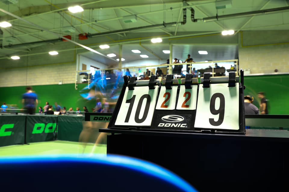 A score board with a close score is in the foreground with the Table Tennis action happening behind it.