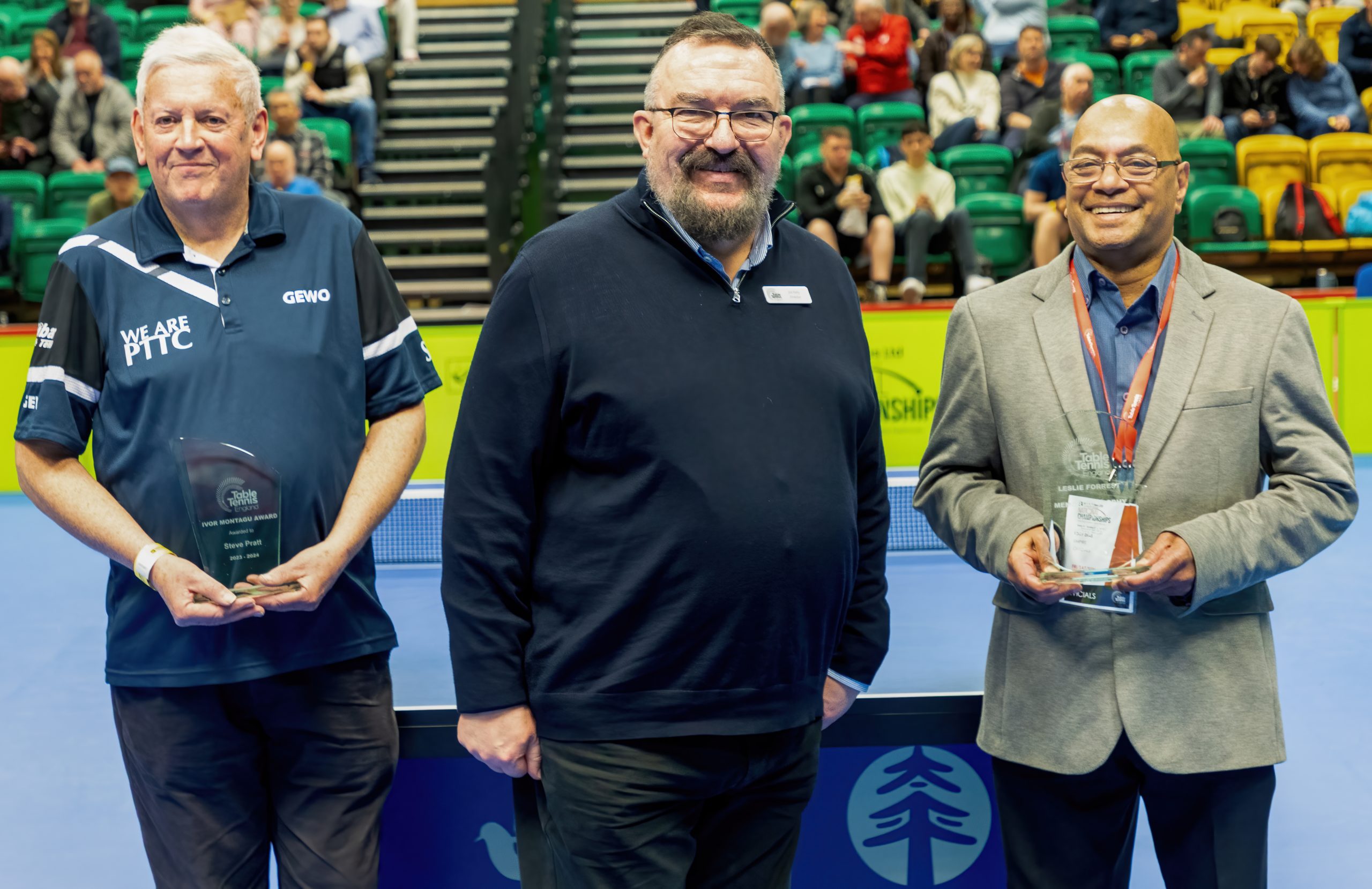 3 men stand in front of a Table Tennis table with a crowd behind them
