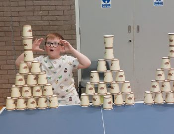 A boy is amazed at his achievement of building a large pyramid of coffee cups as a target on a table tennis table