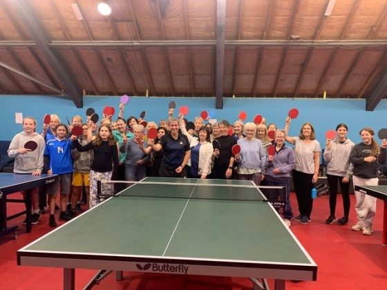 A group of women and girls hold their table tennis bats high, smiling in a group of 30