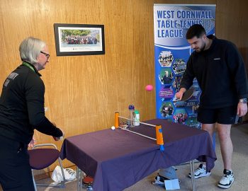 2 people play table tennis around a small table with a banner behind them saying west cornwall table tennis league