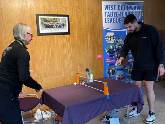 2 people play table tennis around a small table with a banner behind them saying west cornwall table tennis league